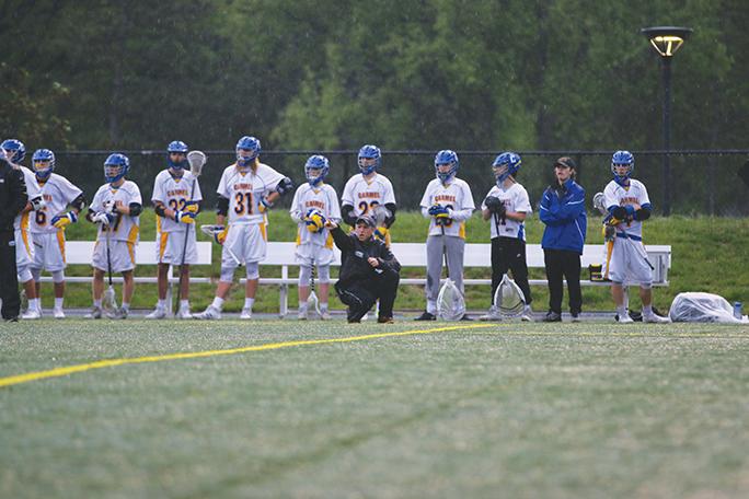 Coach Conway crouches from the sidelines and instructs players on the field while the rest of the team watches. The mens lacrosse team is 10-5-0 for the season.