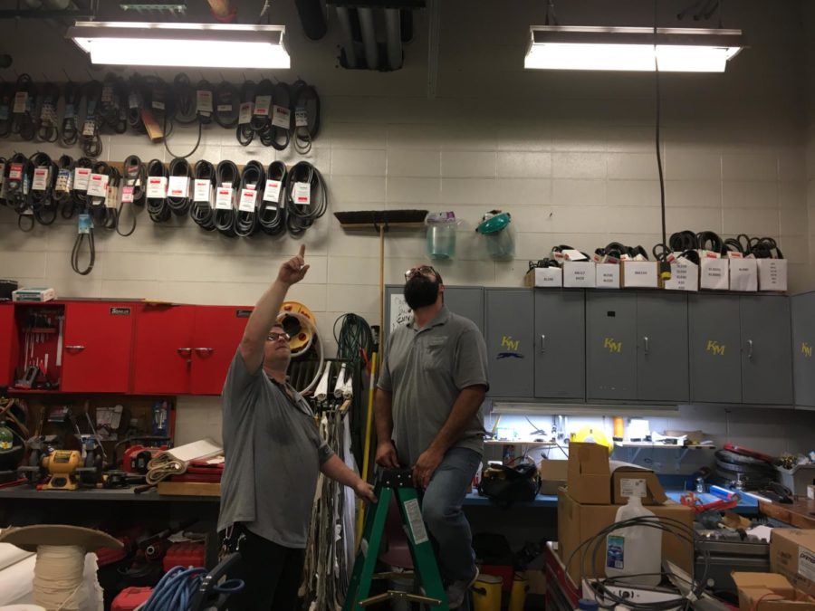 Maintenance worker Fred Napier holds the ladder for another maintenance worker as they fix a light that has gone out in the maintenance shop. This is the same kind of thing they do when teachers submit work orders.