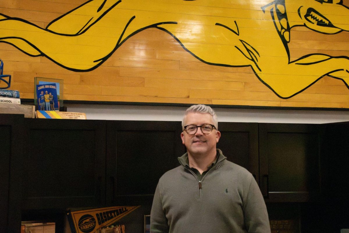 Principal Tim Phares stands in front of his new office wall art made from an old piece of a gym floor. Phares said the administration is hoping to prevent delays during the SAT on March 5 by running a stress test for juniors on Feb. 14. 